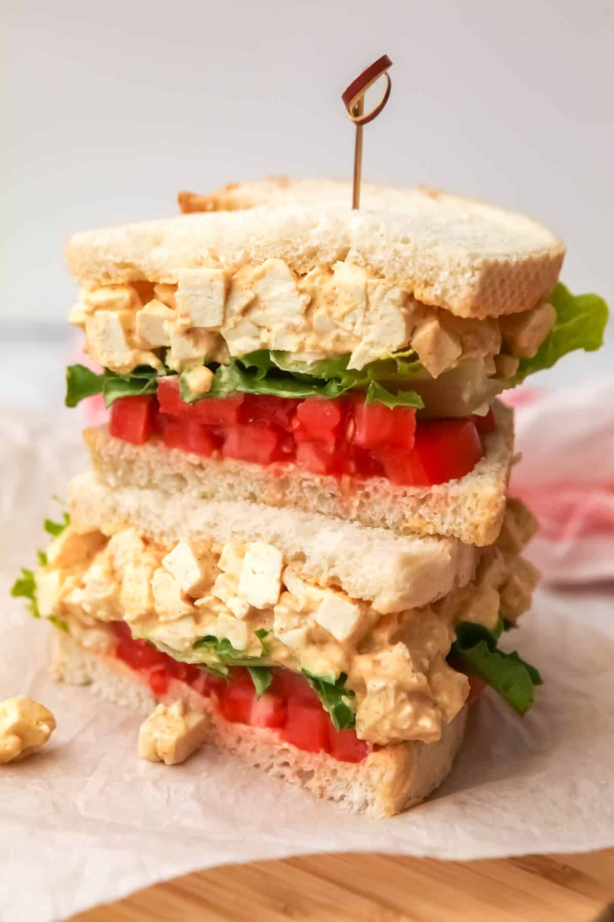 A vegan tofu egg salad sandwich cut in half and stacked on top of eachother on a cutting board.