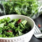 A bowl of kale salad with dried cranberries on top and a larger bowl of salad behind it.