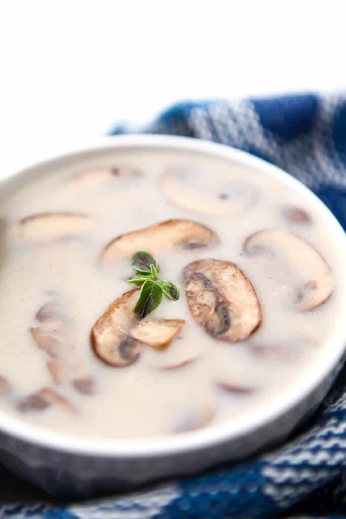 A white bowl filled with cream of mushroom soup with some sliced mushrooms on top with a blue towel around it.