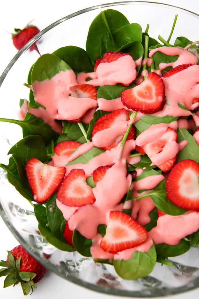 A glass salad bowl filled with spinach and sliced strawberries and drizzled with vegan strawberry vinaigrette dressing.