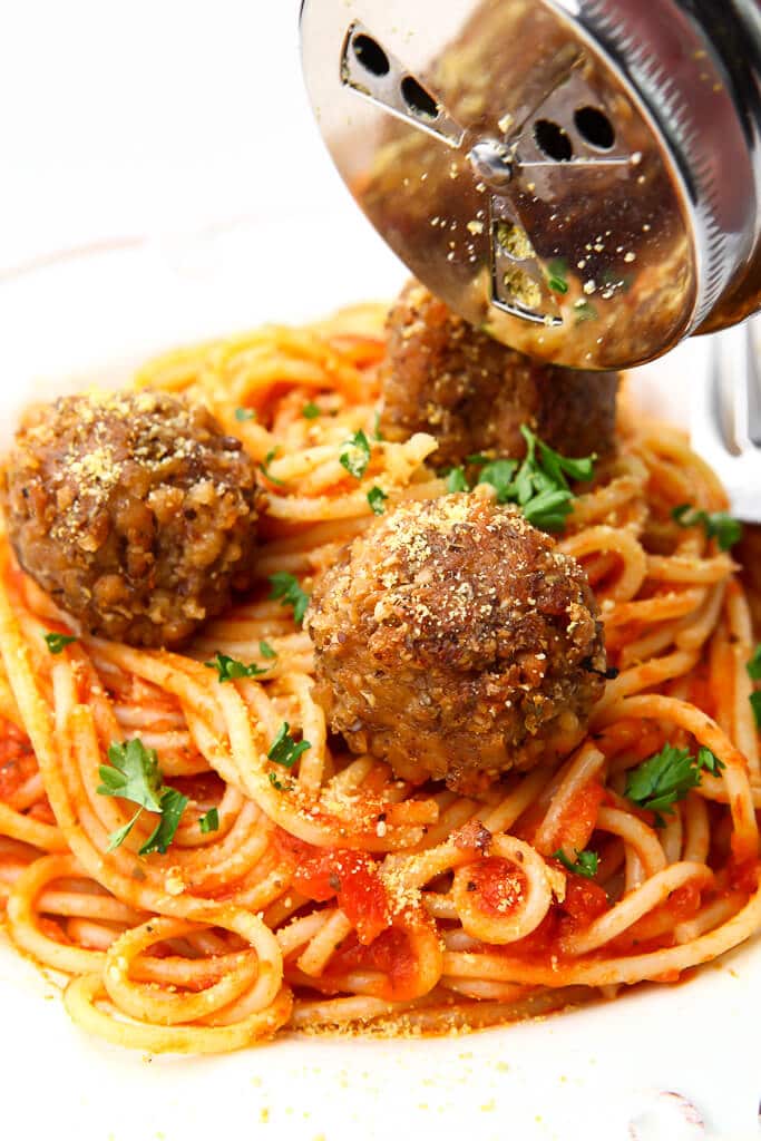 A plate of vegan spaghetti and meatballs with a silver shaker sprinkling vegan Parmesan cheese over the top. 