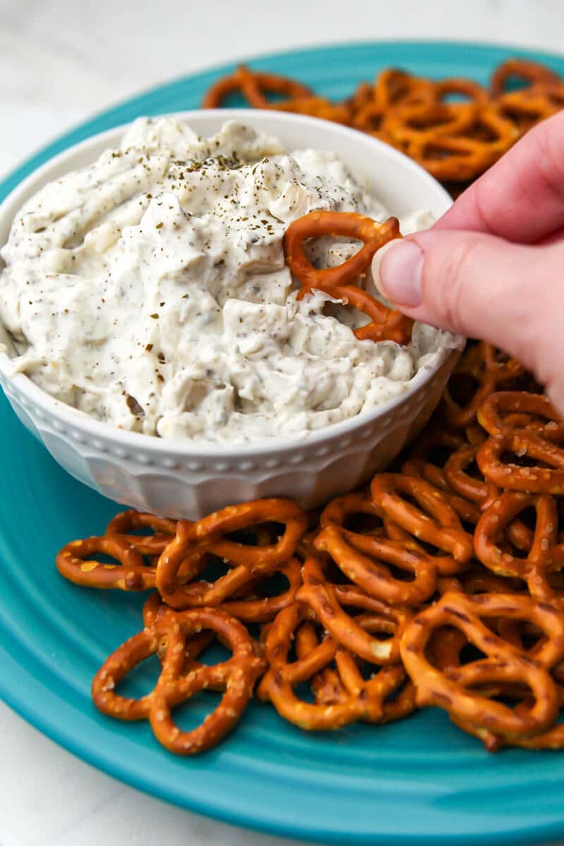 A white bowl filled with mock clam dip and pretzels on the side being scooped up with a pretzel.