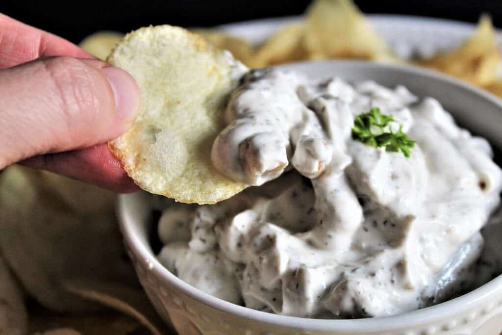 A white bowl filled with vegan clam dip with a chip scooping out some dip.