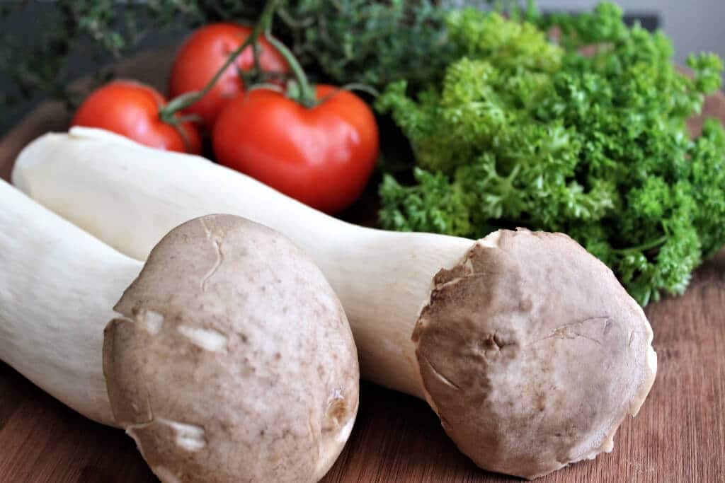 Two king mushrooms on a cutting board with tomatoes and parsley.