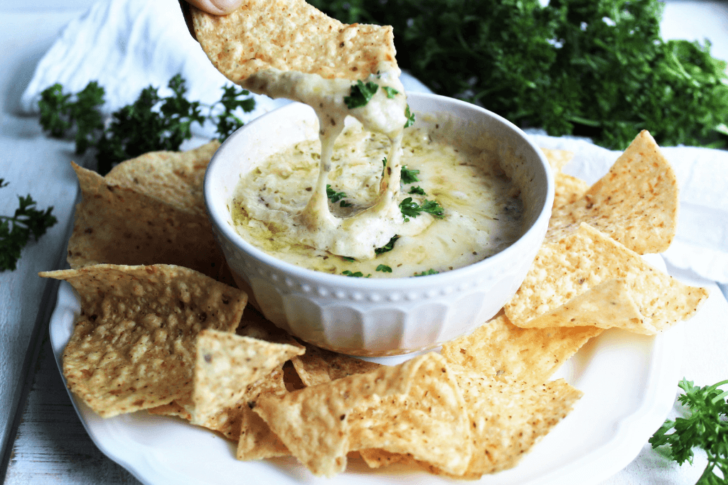 A bowl of easy vegan artichoke dip with chips and parsley around it.