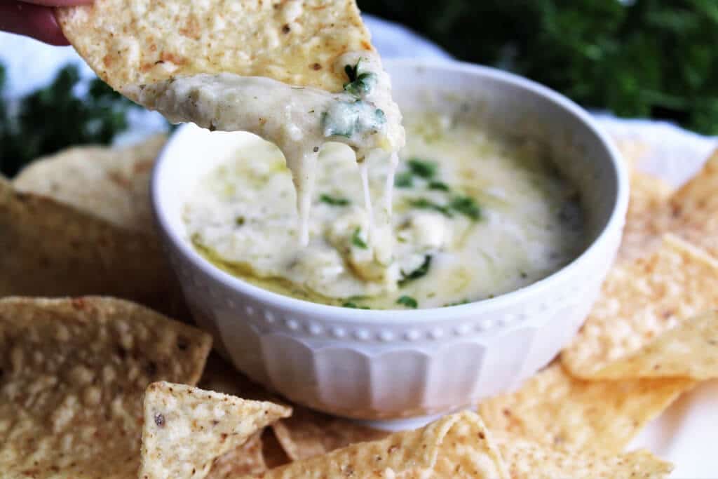 A close up of a chip being dipped into a bowl of artichoke dip with cheese and parsley on top.