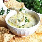 A white bowl filled with easy vegan artichoke dip topped with vegan cheese and parsley.
