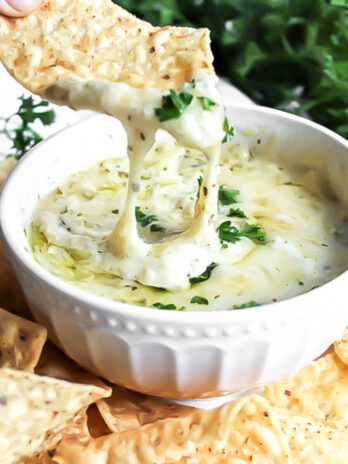 A white bowl filled with easy vegan artichoke dip topped with vegan cheese and parsley.