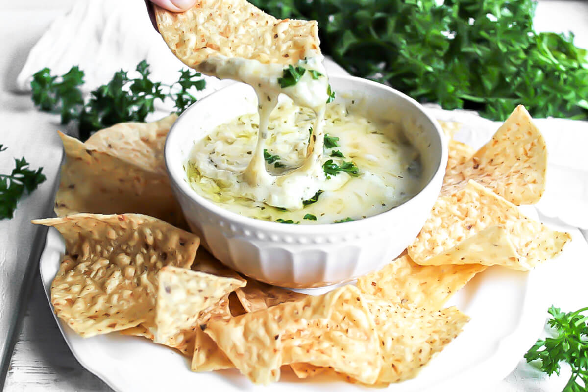 A bowl of easy vegan artichoke dip with chips and parsley around it.