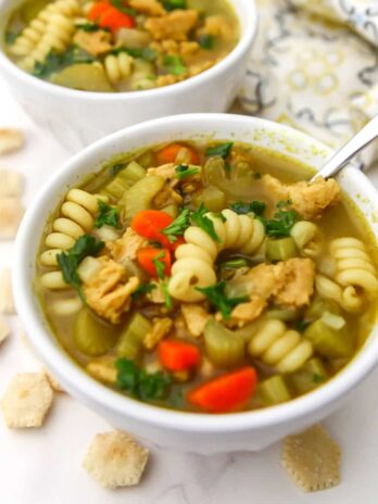 Two bowls of vegan chicken noodle soup made with soy curls with soup crackers around them.