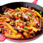 A red iron skillet filled with peppers, onions, portabella mushrooms, and soy curl "chicken" strips to make vegan fajitas..