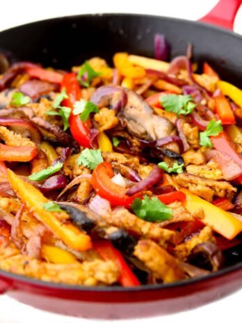 A red iron skillet filled with peppers, onions, portabella mushrooms, and soy curl "chicken" strips to make vegan fajitas..