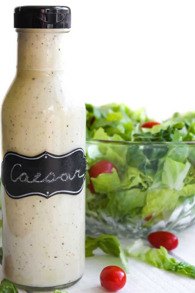 A glass jar filled with dairy-free Caesar dressing with a bowl of salad behind it. 