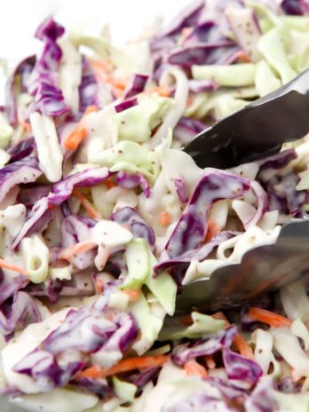 A close up of vegan coleslaw in a bowl with tongs scooping some out.
