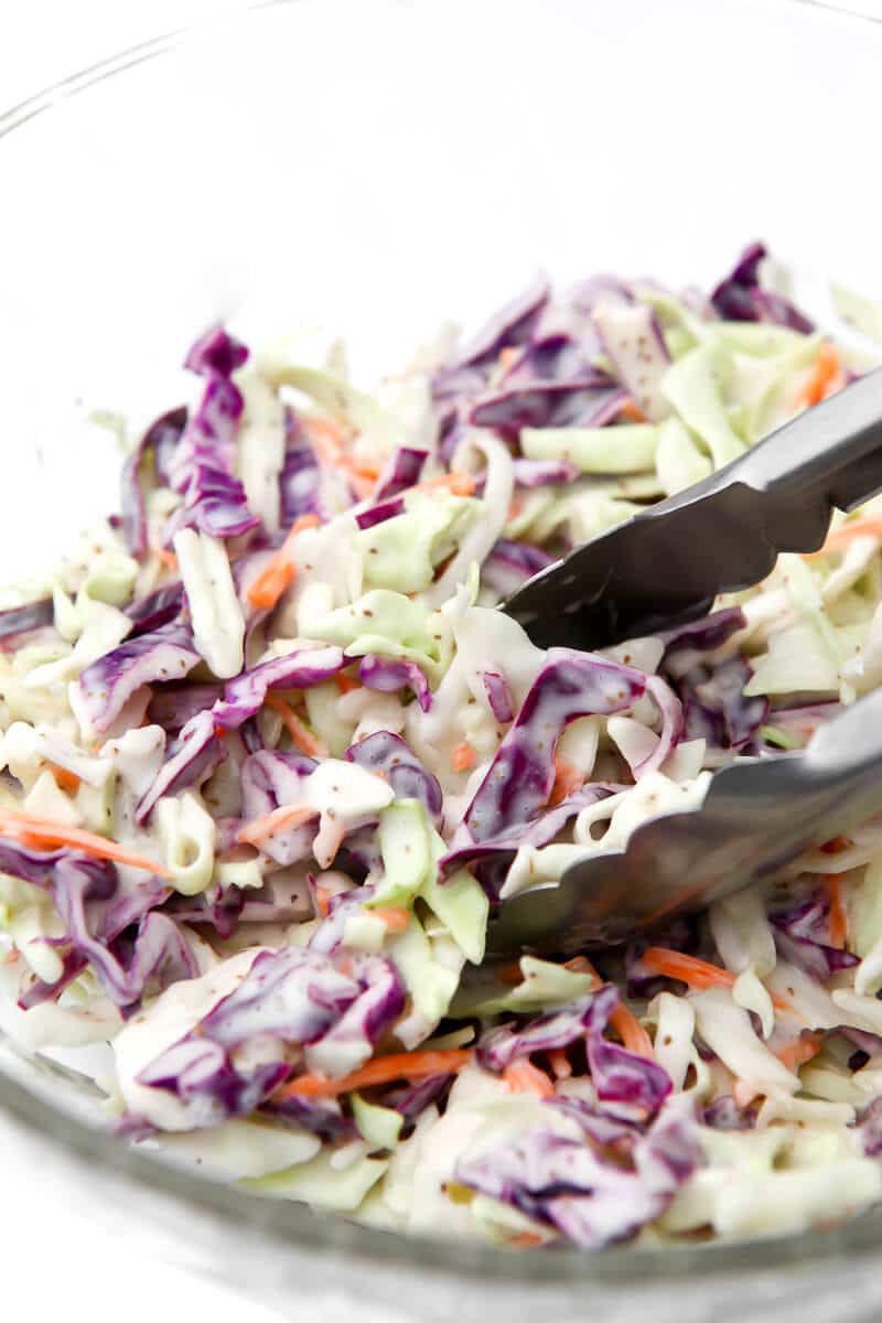 A glass bowl filled with creamy coleslaw with tongs scooping some out.