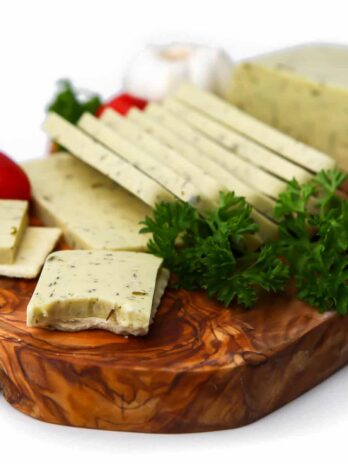 A block of vegan garlic herb cheese on a wooden cutting board with herbs and cherry tomatoes around it.