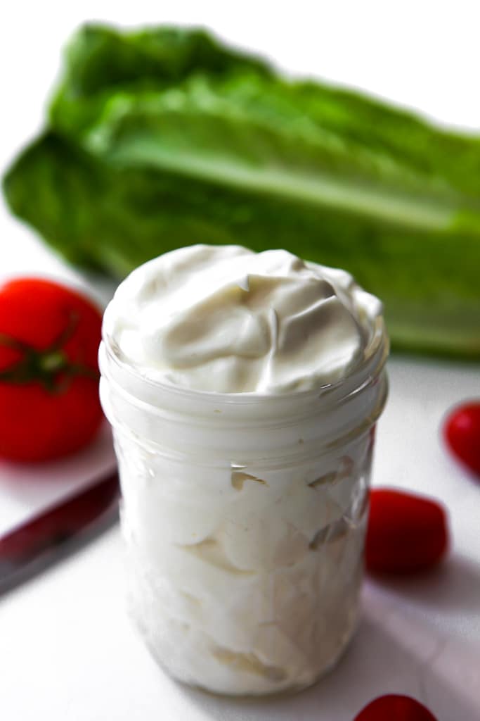 A mason jar filled with vegan mayo made with soy milk and oil with some lettuce and tomatoes around it.