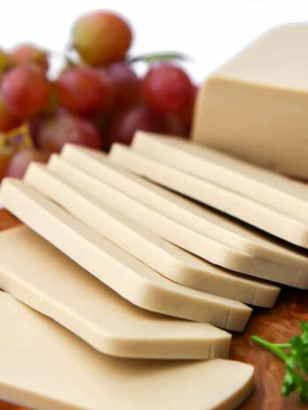 A close up of vegan smoked Gouda cheese sliced with grapes behind it.