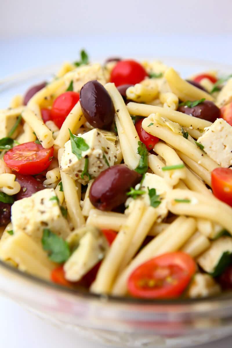 A glass bowl filled with vegan Greek pasta salad with vegan tofu feta, tomatoes, olives and herbs.