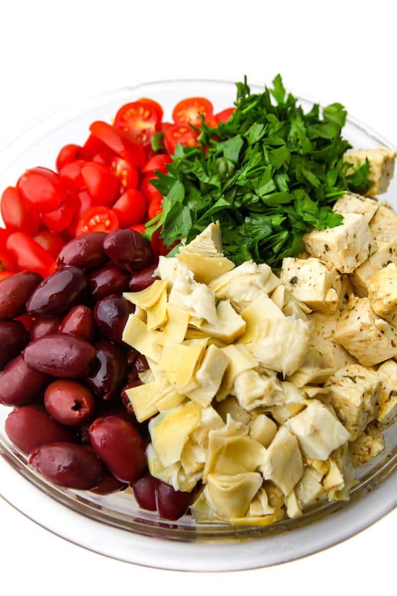A bowl filled with halved cherry tomatoes, chopped parsley, kalamata olives, artichoke hearts, and vegan feta to make a vegan Greek salad.