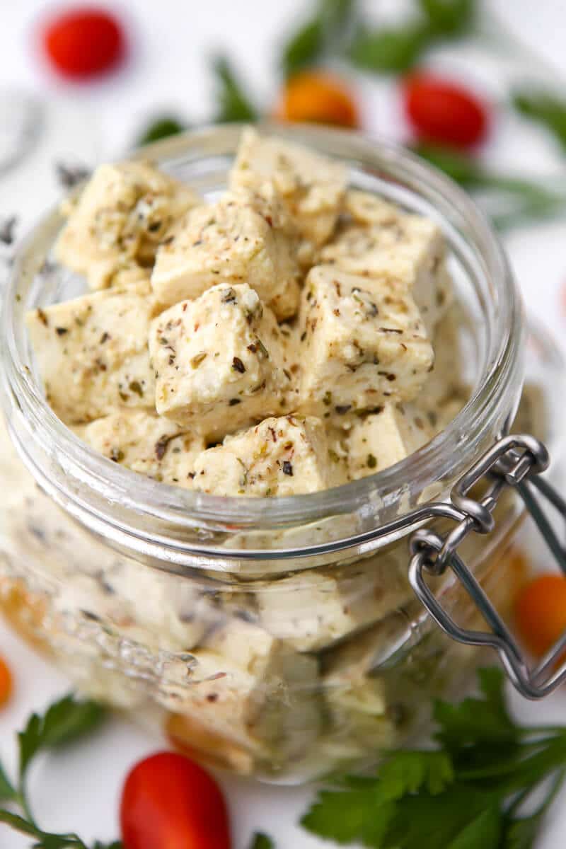 A jar of vegan feta cheese made from tofu with cherry tomatoes and parsley around it.