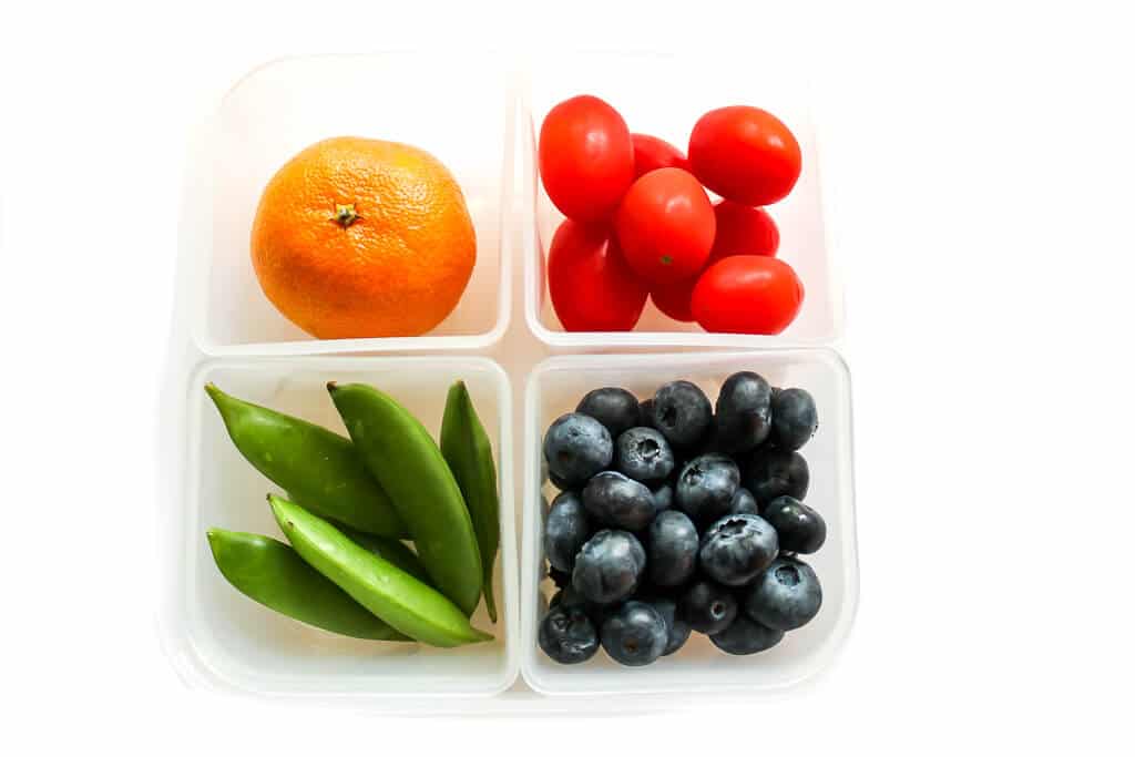 A container with 4 compartments filled with an orange, snap peas, blueberries, and cherry tomatoes for kids lunches.