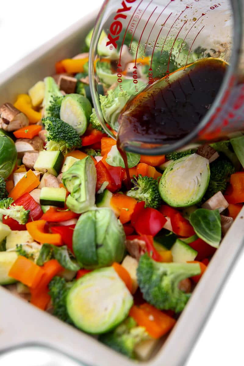 A baking dish filled with assorted vegetables being drizzled with maple balsamic dressing.