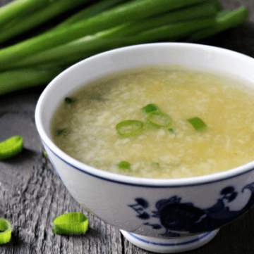 A close up of vegan egg drop soup in a white bowl with green onions in the background.
