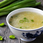 A close up of vegan egg drop soup in a white bowl with green onions in the background.