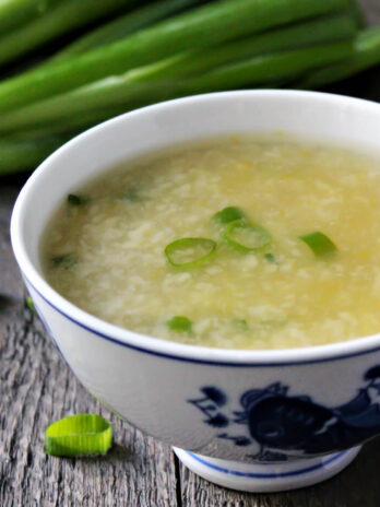 A close up of vegan egg drop soup in a white bowl with green onions in the background.