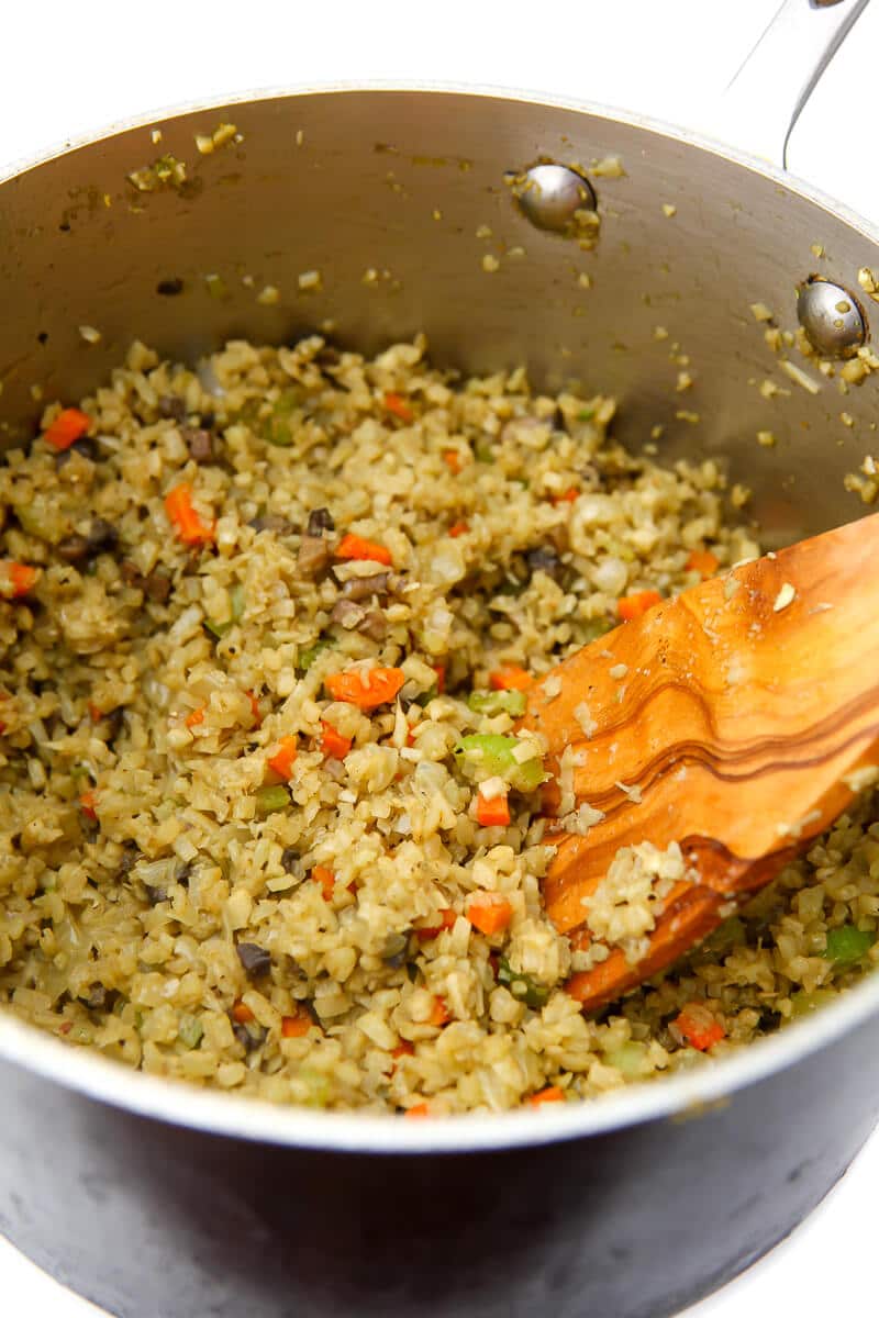 Riced cauliflower and poltry seasoning added to sauted veggies in a pan being stirred with a wooden spoon.
