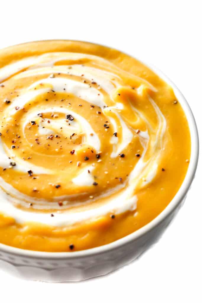 A close up of a white bowl filled with creamy vegan butternut squash soup with some vegan sour cream and ground pepper swirled on top.