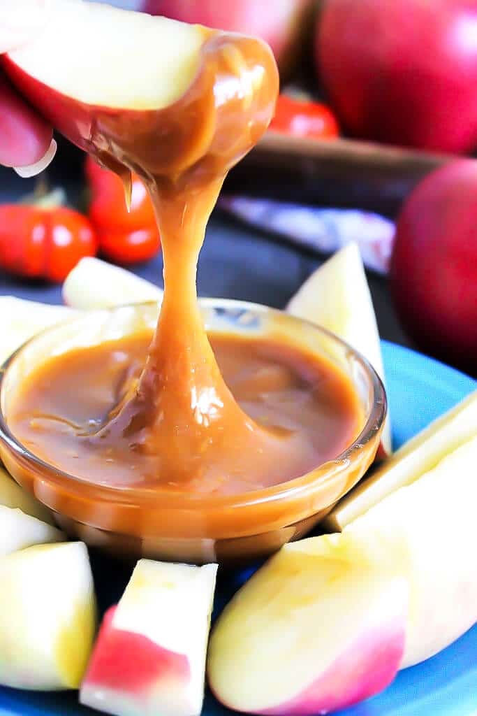 An apple slice being dipped into a bowl of vegan salted caramel sauce.
