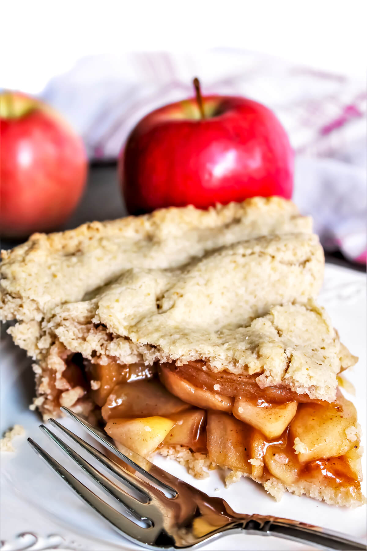 A slice of gluten-free vegan apple pie with 2 apples behind it and a fork on the side.
