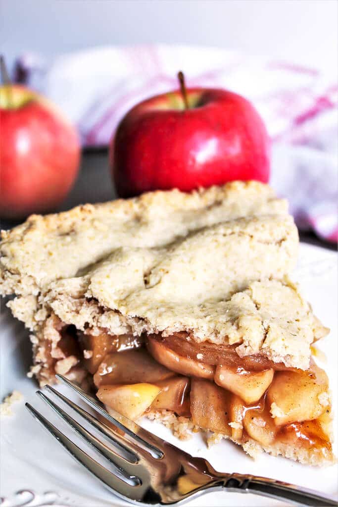 A slice of gluten-free vegan apple pie with 2 apples behind it and a fork on the side.