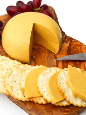A round of vegan cheddar cheese cut and sliced on crackers on a wooden cutting board.