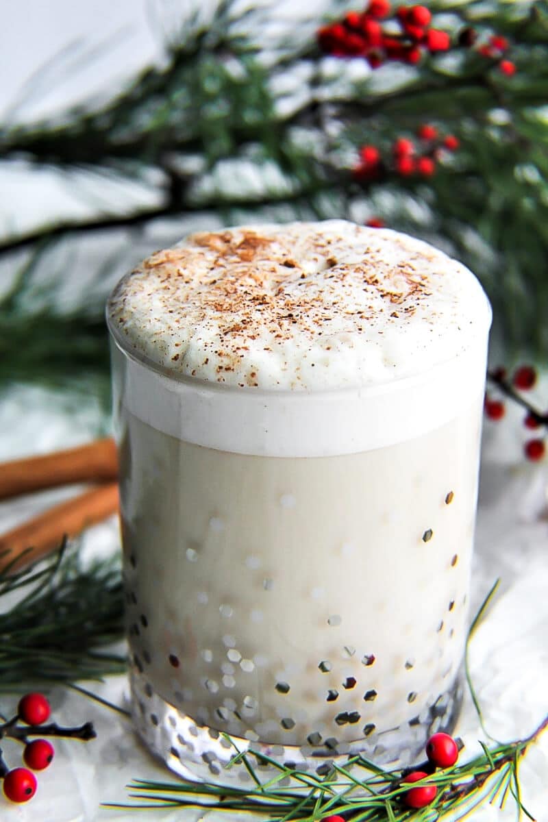 A glass of homemade vegan eggnog with cinnamon sticks, pine boughs and red berries around it.