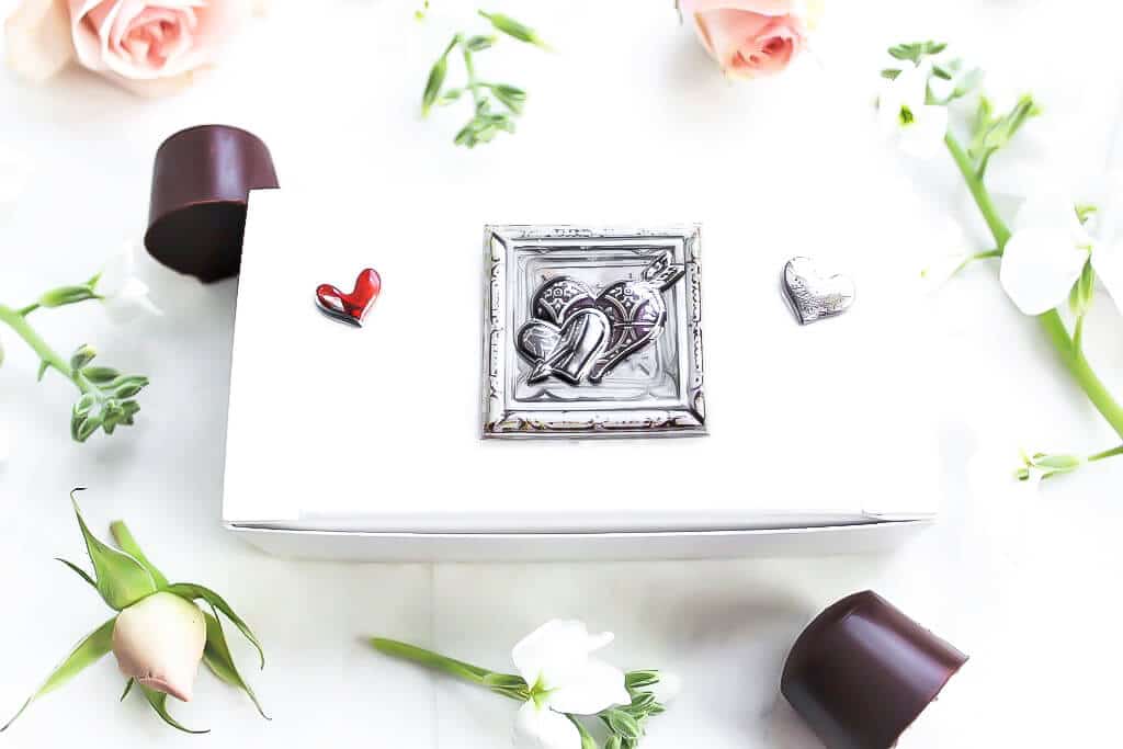 A white candy box filled with chocolates with flowers around it. 