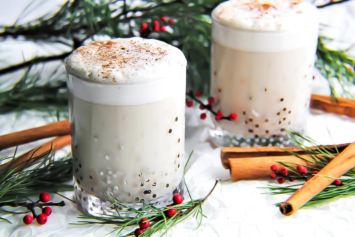 Two glasses of vegan eggnog with cinnamon sticks and red berries around them.