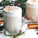 Two glasses of vegan eggnog with cinnamon sticks and red berries around them.