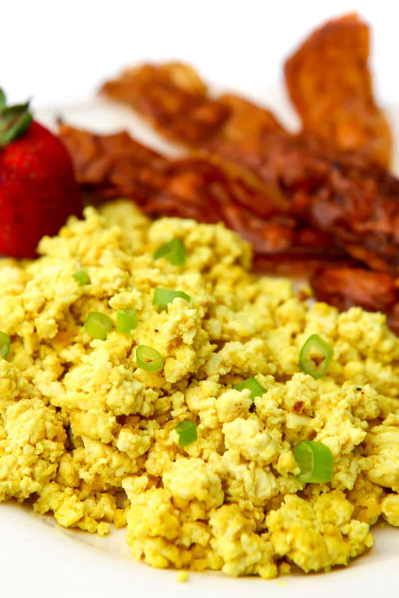 Vegan scrambled eggs made from tofu topped with green onions with vegan bacon and a strawberry on the side.
