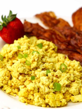 Tofu scramble made to taste like eggs topped with green onions with vegan bacon and a strawberry on the side.