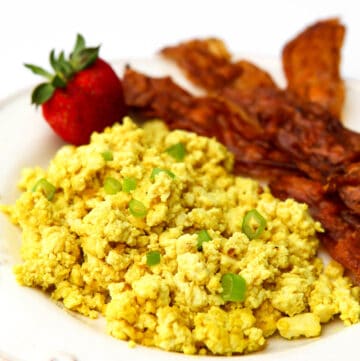 Tofu scramble made to taste like eggs topped with green onions with vegan bacon and a strawberry on the side.