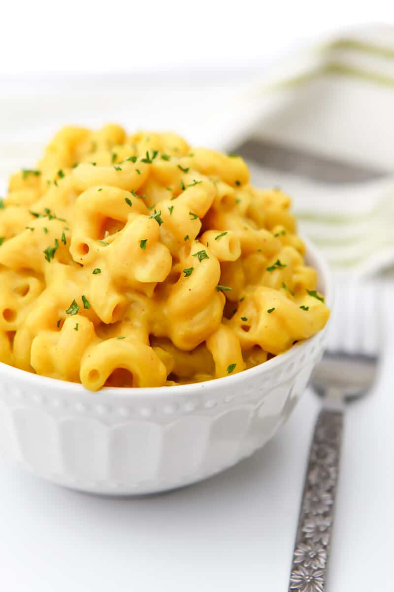 A bowl of vegan macaroni and cheese with a green and gray tea towel behind it and a fork next to it.