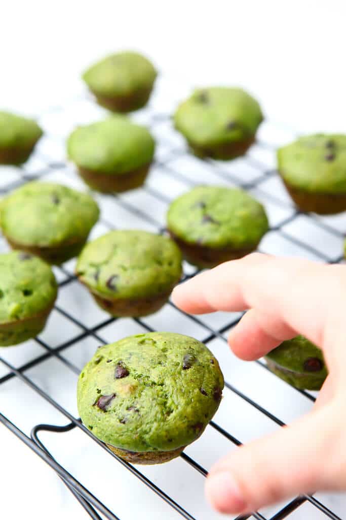 Green mint chocolate chip muffins on a cooling rack with a kid's hand reaching for one.