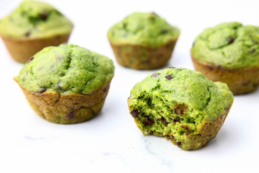 Five green spinach muffins on a marble counter top with a bite taken out of them 