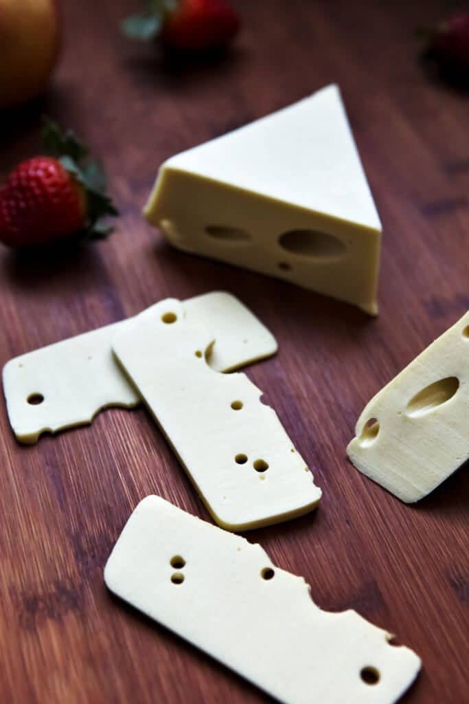A triangle shaped block of vegan Swiss cheese with slices of cheese and strawberries around it on a wooden cutting board..