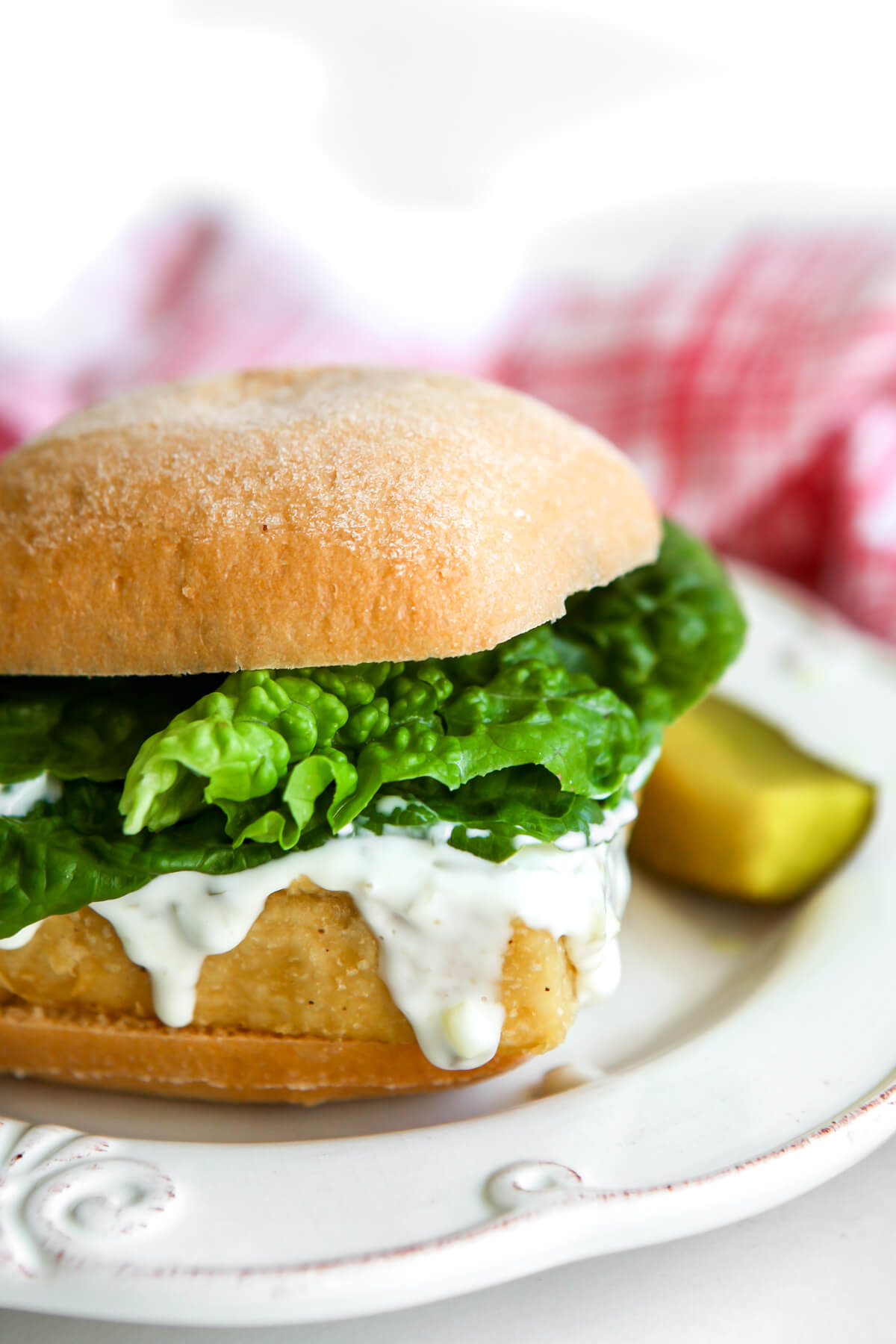 A tofish sandwich with tartar sauce and lettuce and a pickle on the side.