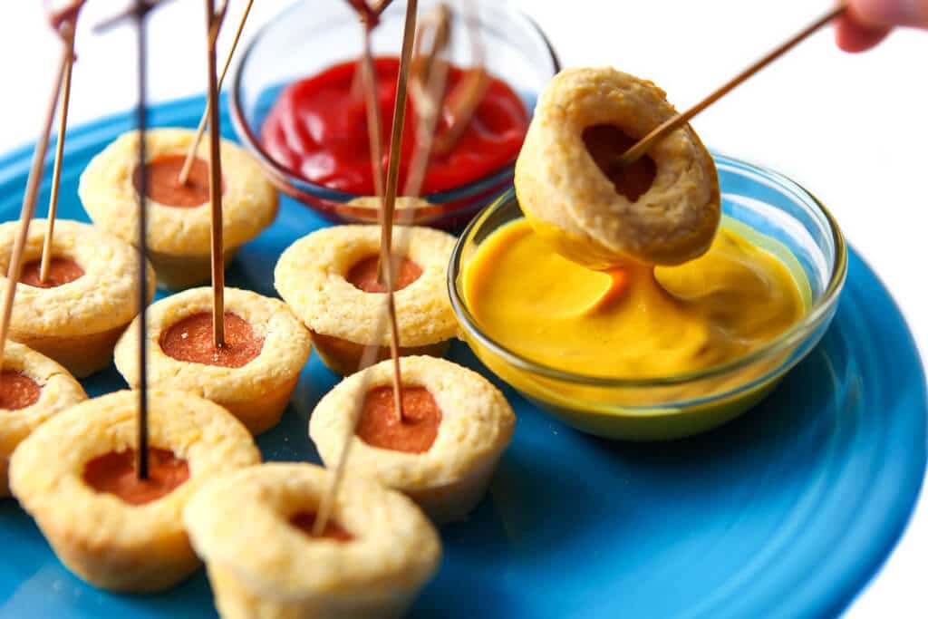 A blue plate filled with mini vegan corn dog bites with one being dipped into a bowl of mustard.