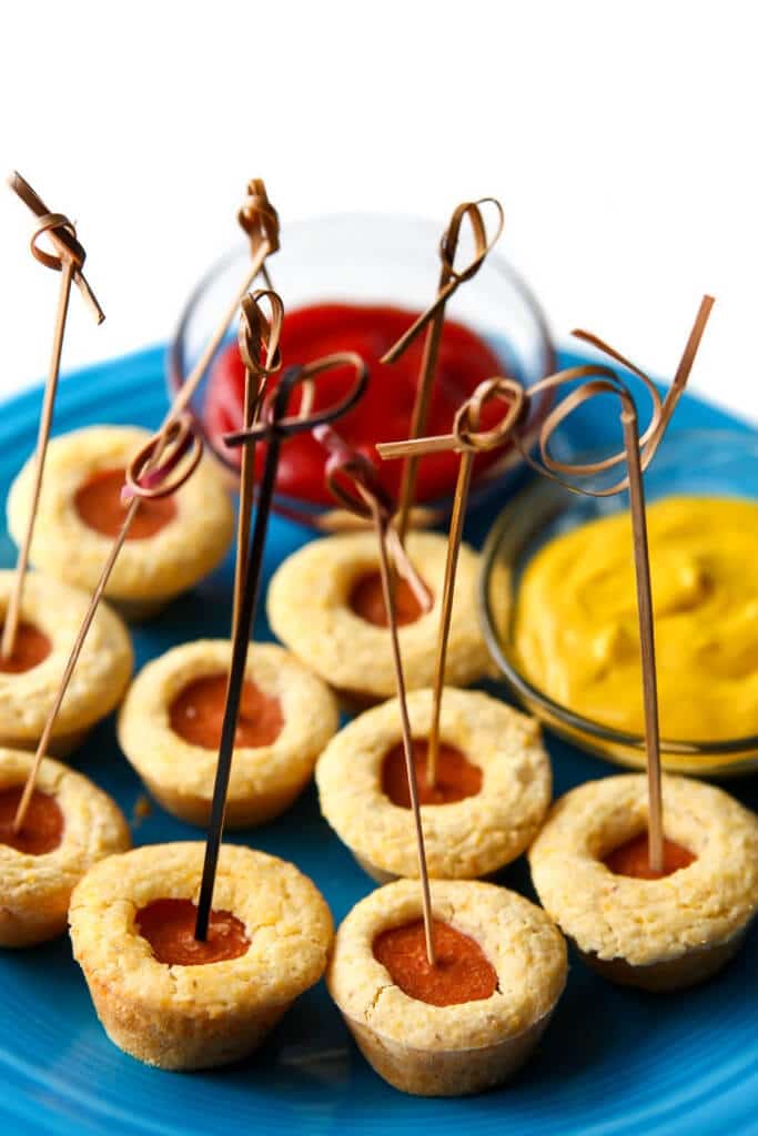 A blue plate filled with mini vegan corn dog bites with small bowls of ketchup and mustard for dipping on the side.